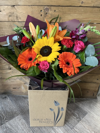 Sunburst Garden - A vibrant, hand-tied bouquet bursting with warm, cheerful energy. Bold golden tones, oranges and pops of pink and deep red blooms. Accented with fresh greenery and soft eucalyptus leaves, the arrangement feels both rustic and refined. Wrapped paper and presented in a kraft box, this bouquet evokes the feeling of a bright summer day—perfect for celebrating joy, warmth, and heartfelt moments.